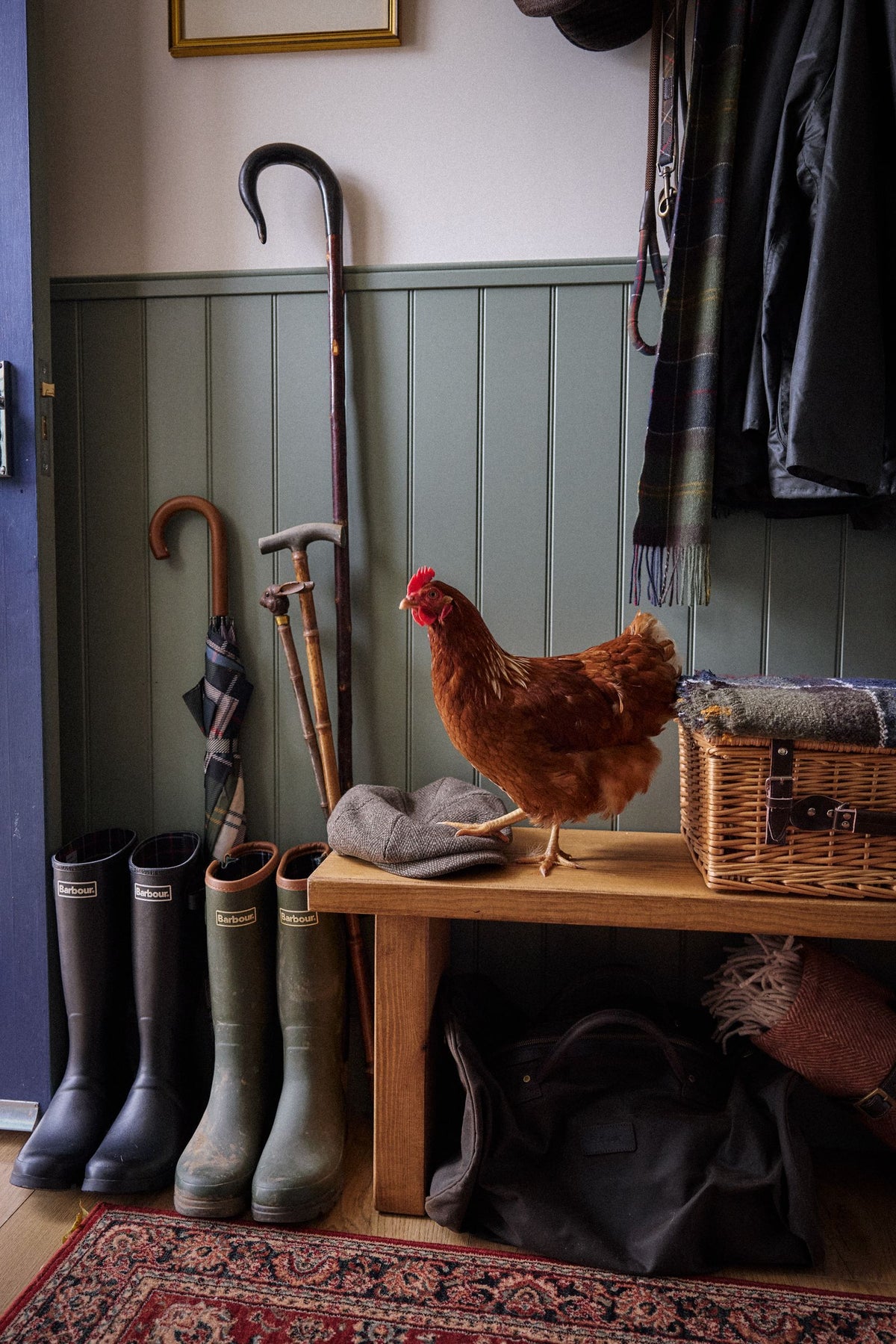 A country boot room with Barbour wellingtons, walking sticks, a flat cap and a hen on the bench