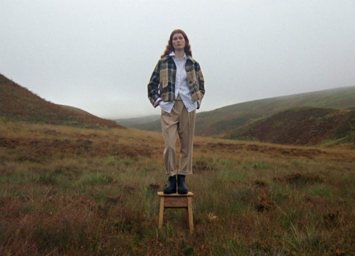 A woman in a Barbour tartan jacket standing on a stool on a Highland moor