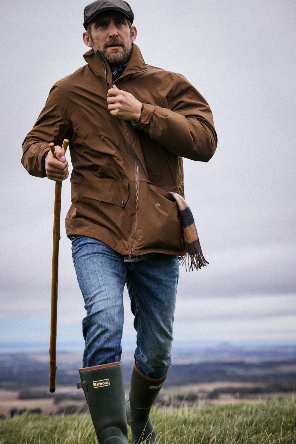 Man in a Barbour waterproof jacket and wellingtons striding up a hill with a walking cane