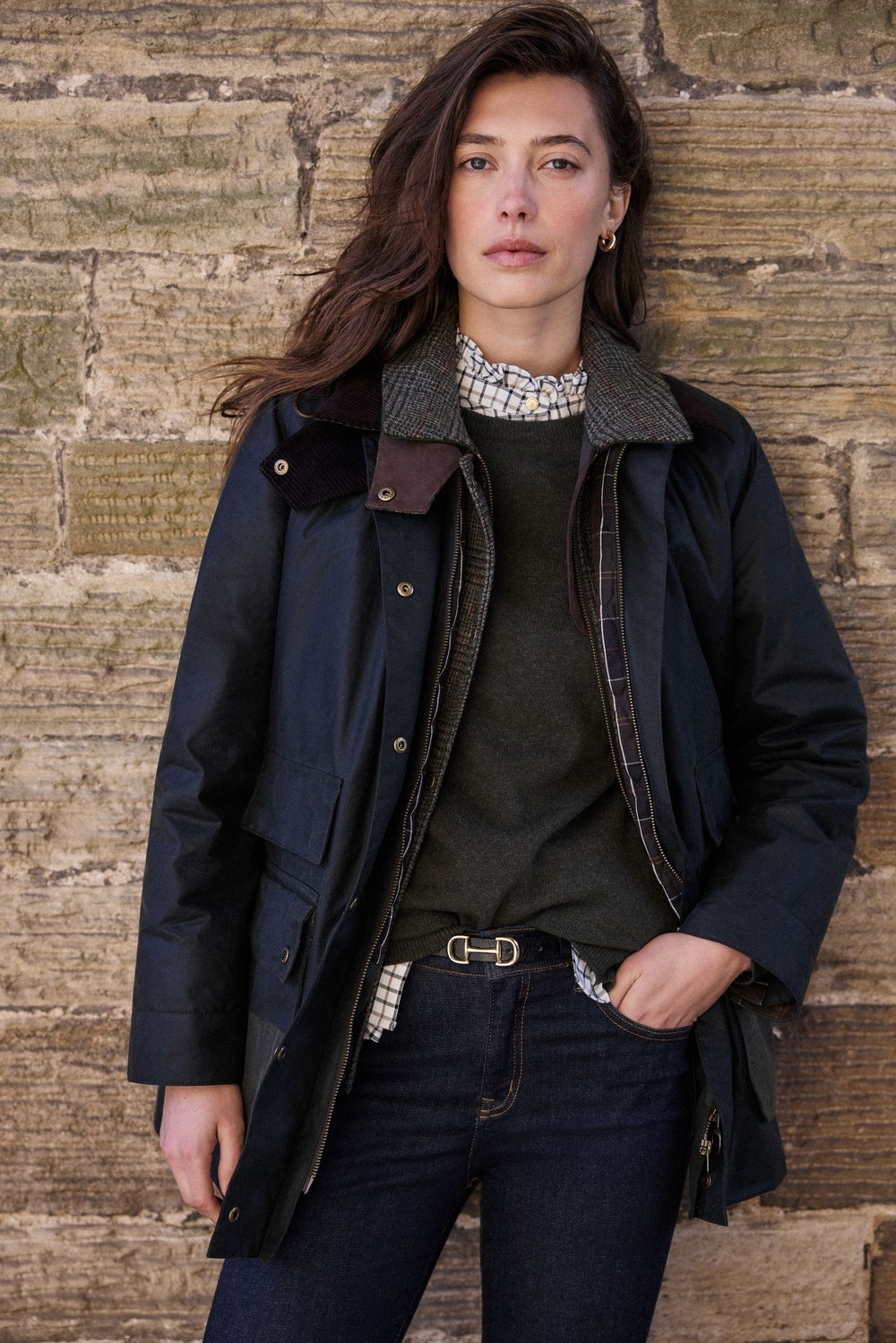 Woman in a navy Barbour waxed jacket leaning against a sandstone wall