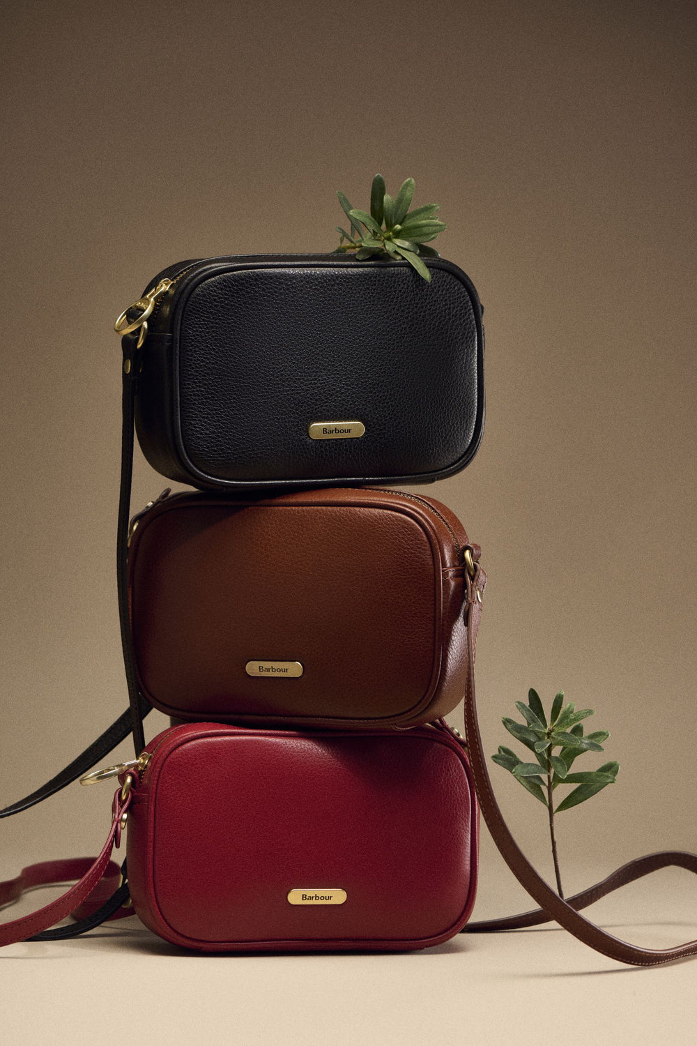 Three Barbour leather crossbody bags stacked with sprigs of greenery